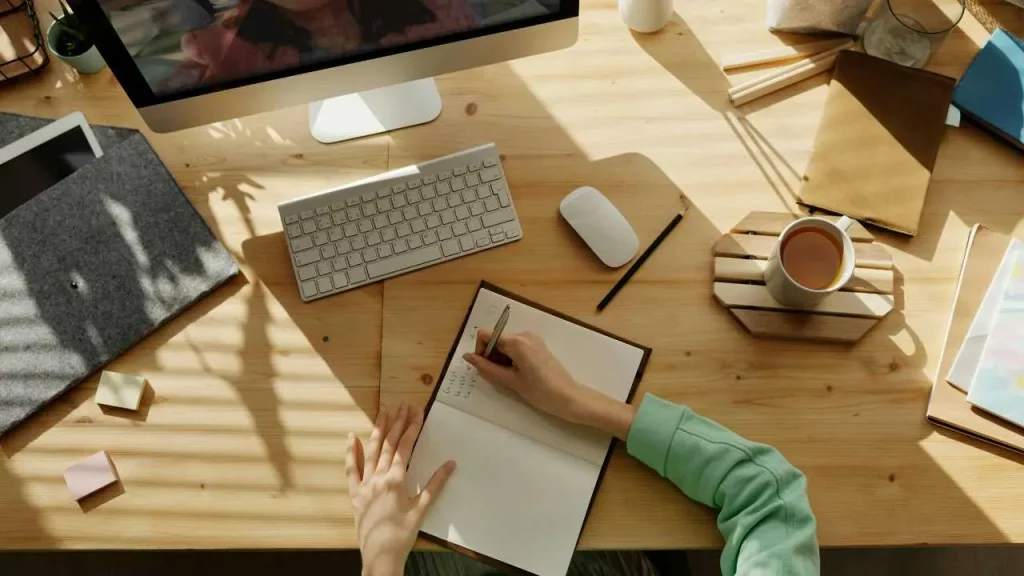 A person's hand writing on a notebook on a table making plans. A cup of office beside, with pencils, mouse, papers scattered on the table. 