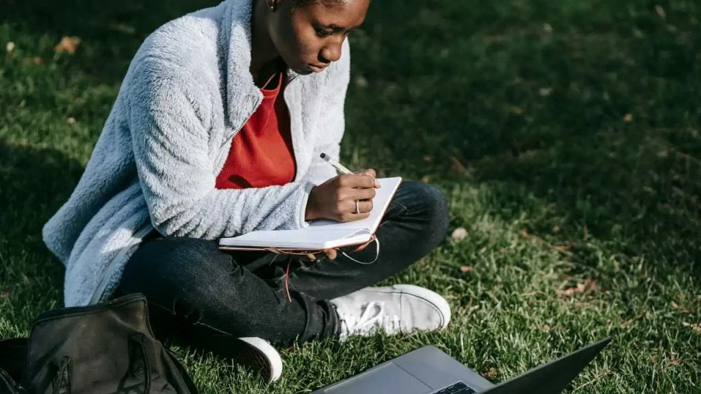 A black woman writing while sitting on a green grass representing how to stop procrastinating