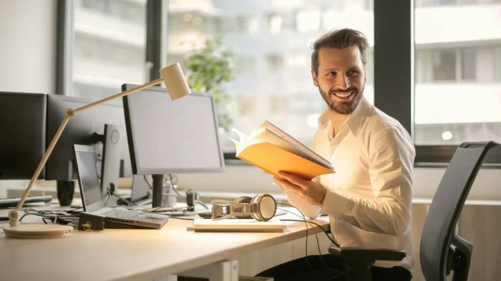 A man holding a yellow note book and sitting in an organized work space