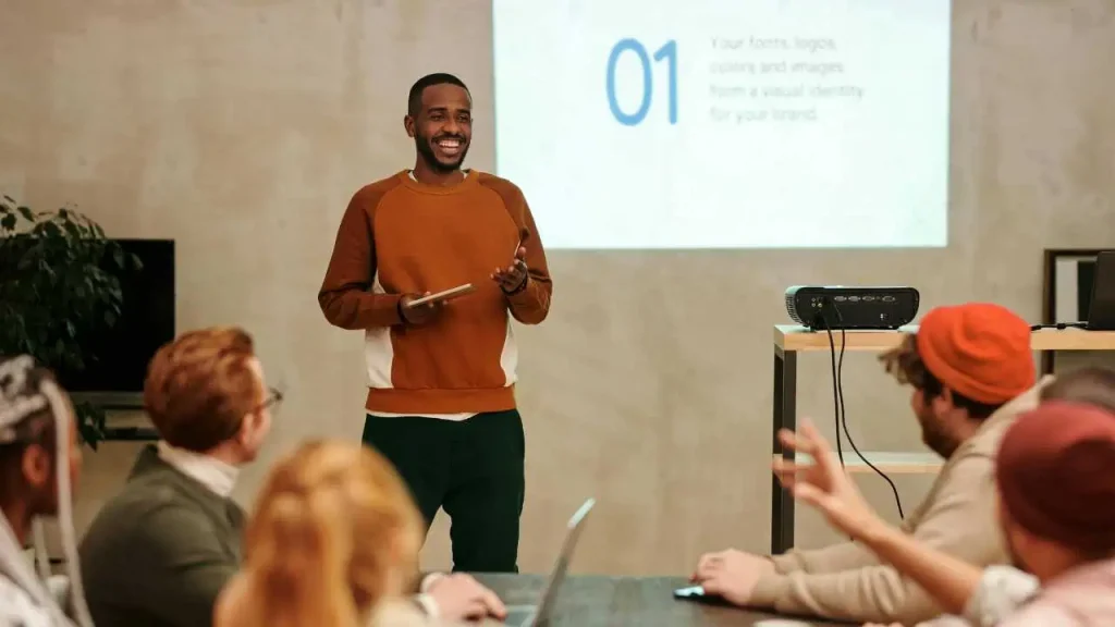 A man in brown shirt presenting a slide to a group of persons.
