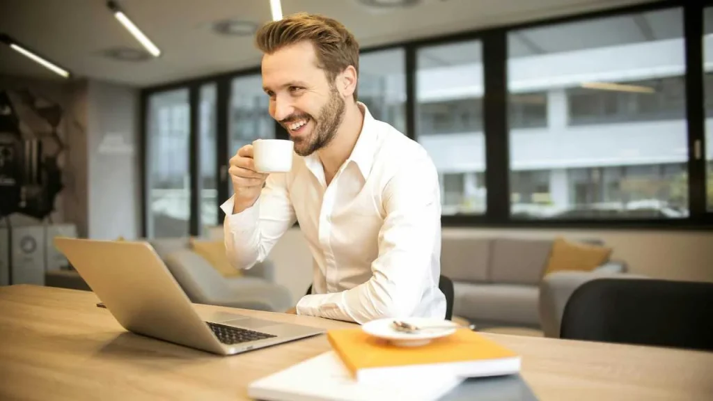 A man on white sleeves happily sipping coffee from a mug while typing his computer